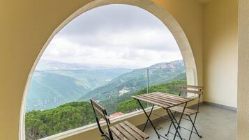 A balcony with a wooden table and chairs, a glass railing, and a view of a mountainous landscape.