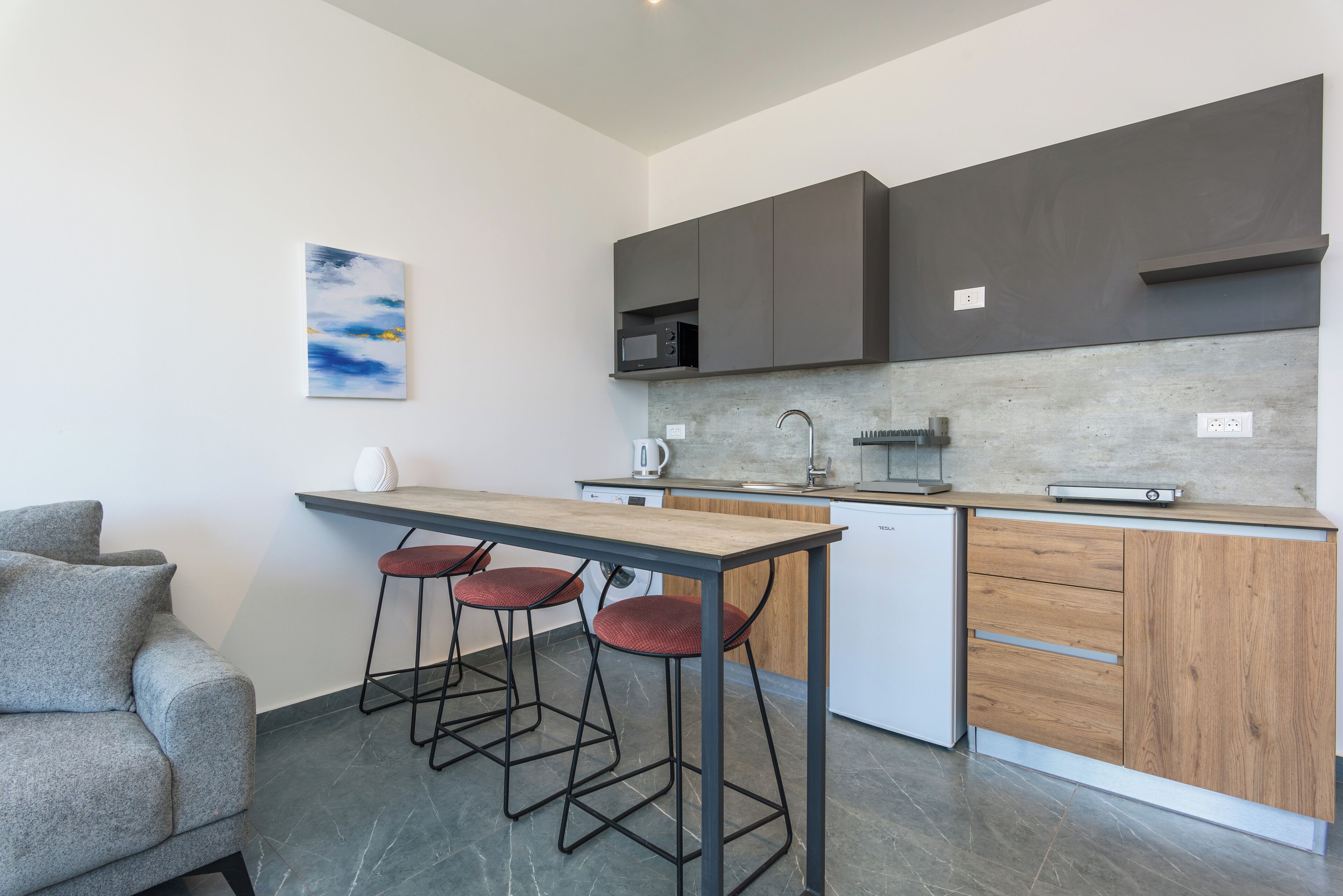 A modern kitchen with a wooden countertop, bar stools, and a grey sofa.