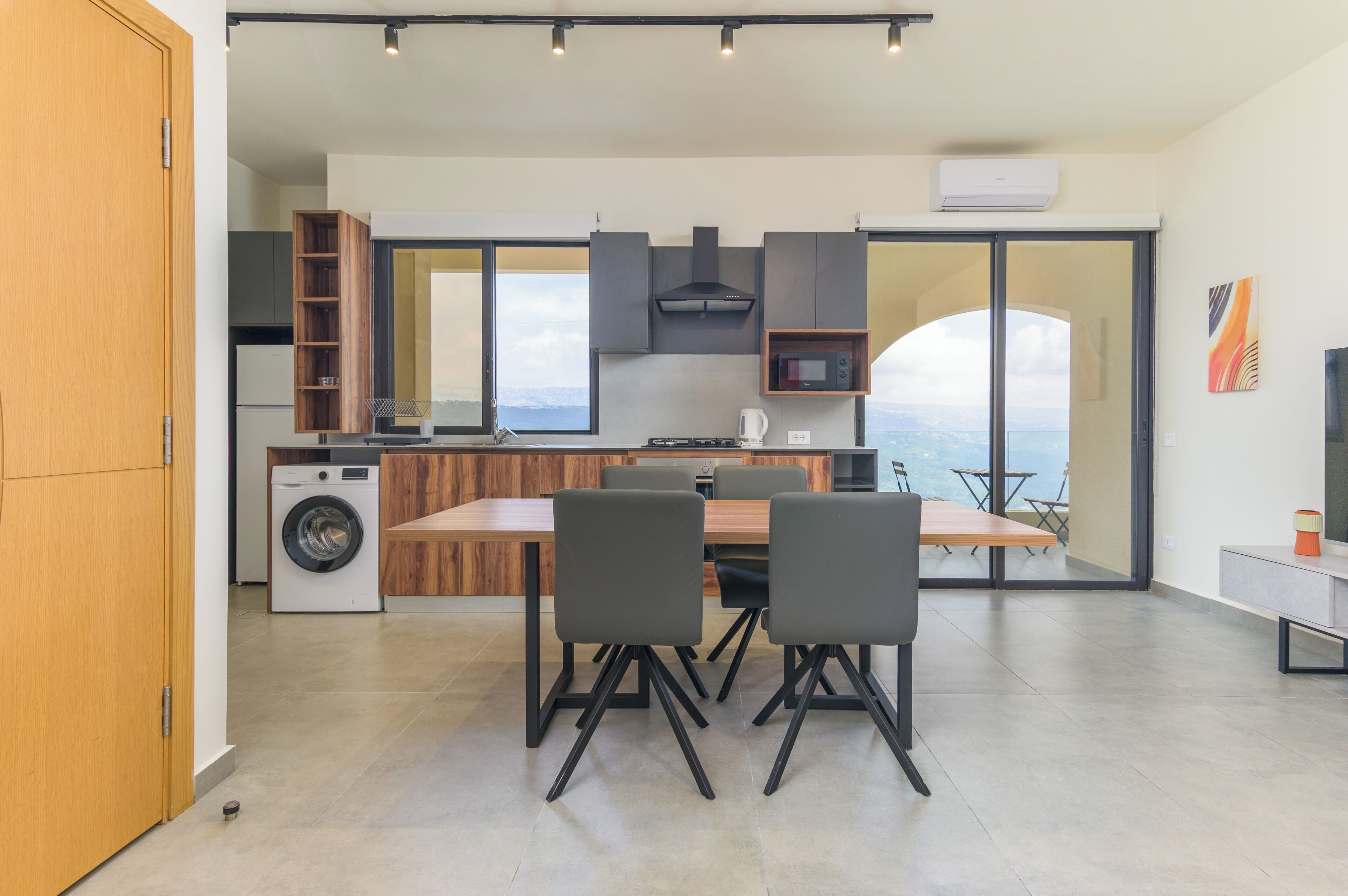 A modern kitchen with a built-in washer, a dining area with a table and chairs, and a large window offering a view of the sea.