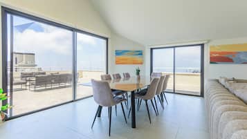 A spacious dining area with a large table and chairs, large windows with a view of the sea, and a balcony with outdoor seating.