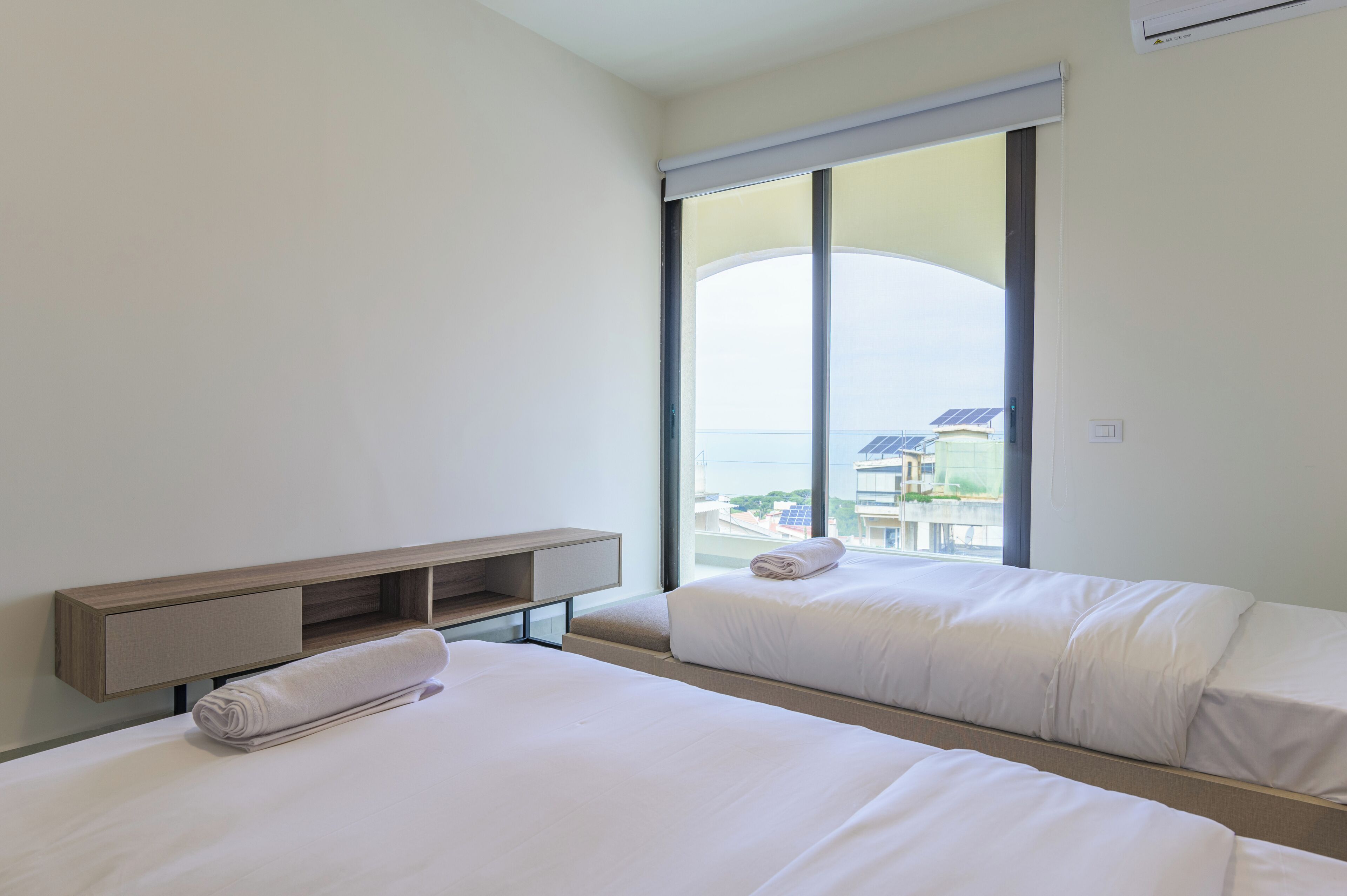 A hotel room with two beds, a wooden bedside table, and a large window offering a view of the city.