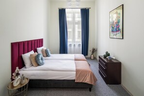 Uma cama de casal com cabeceira vermelha, duas mesas de cabeceira, um penteou e uma janela com cortinas azuis, além de um quadro na parede.