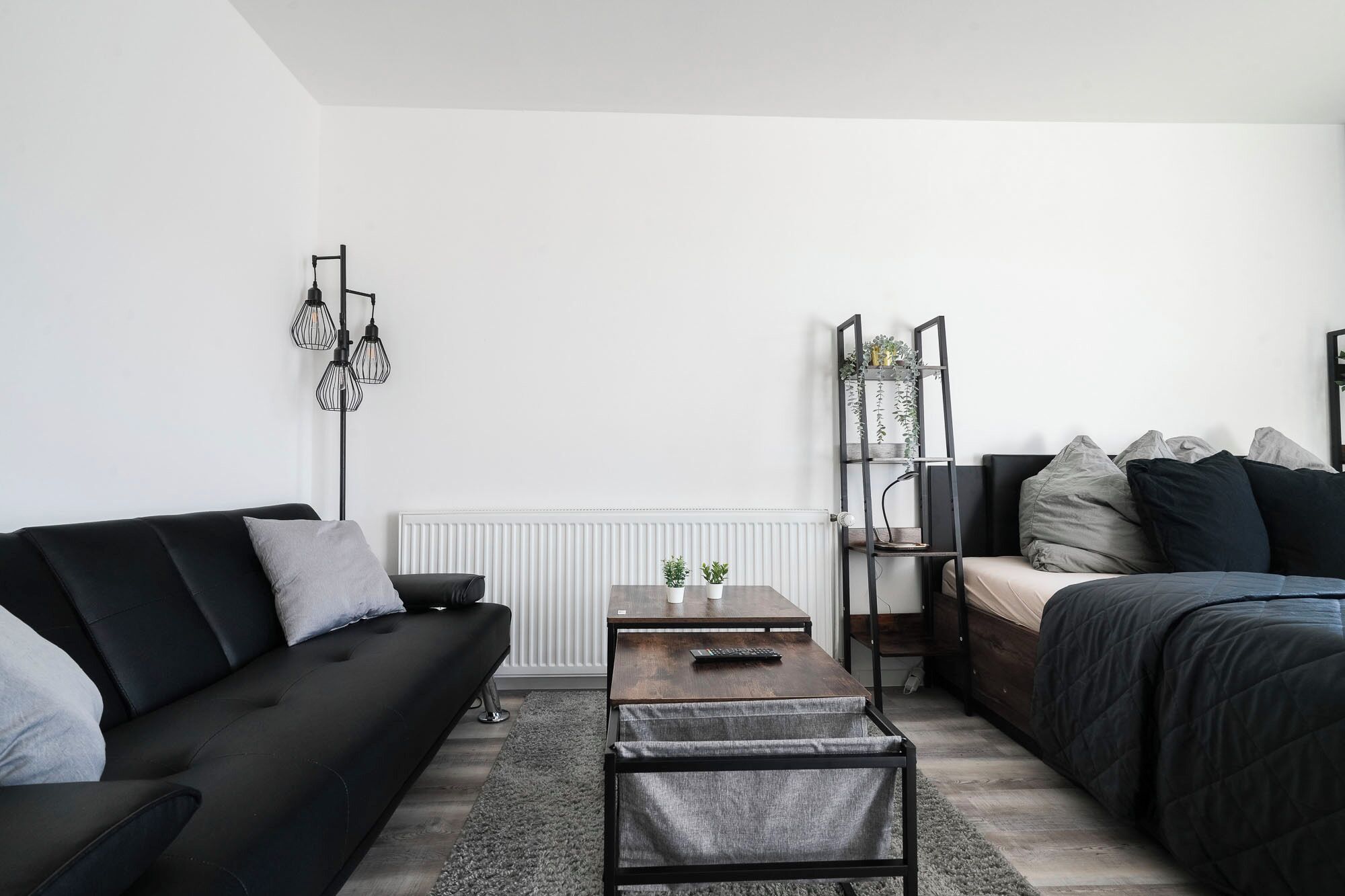 Un soggiorno moderno con un divano nero, un tavolino in legno, un letto con copripiumino scuro, una lampada da terra nera e un termosifone bianco.