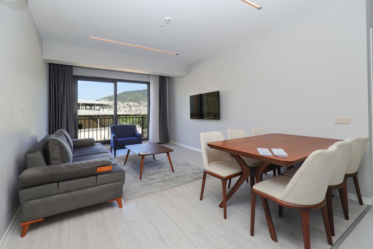 A modern living room with a sofa, a coffee table, and a dining table with chairs. There is a large window with a view of a cityscape and hills.