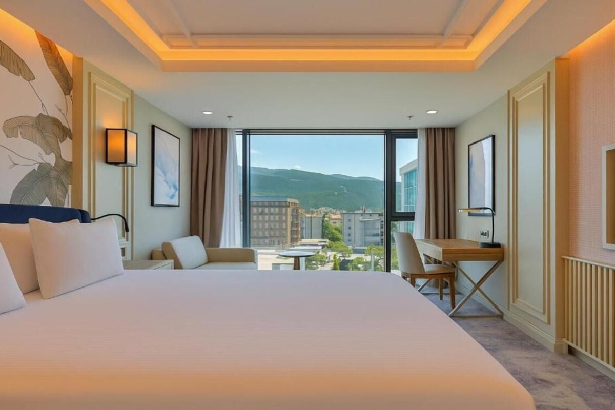 A hotel room with a large bed, a desk, and a chair, a large window with a view of buildings and mountains, and a television on the wall.