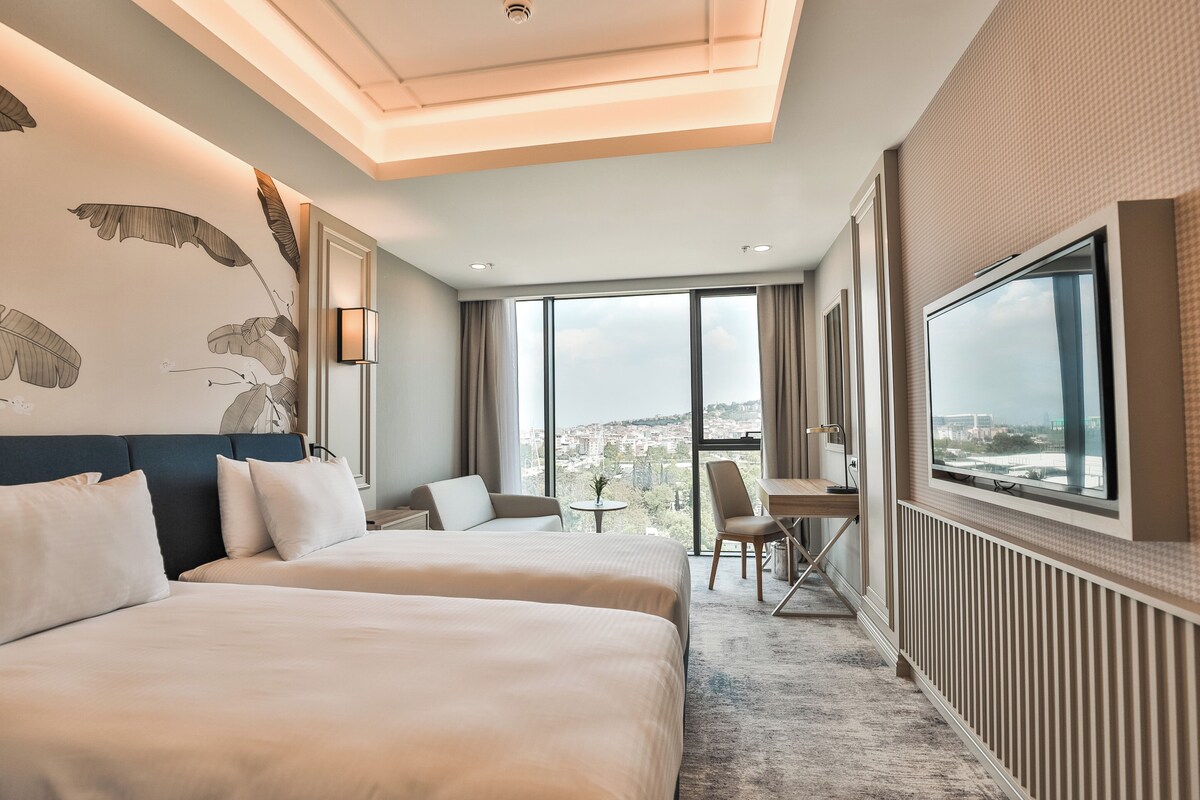 A hotel room with two beds, a desk, a chair, a large window with a city view, and a ceiling light fixture.