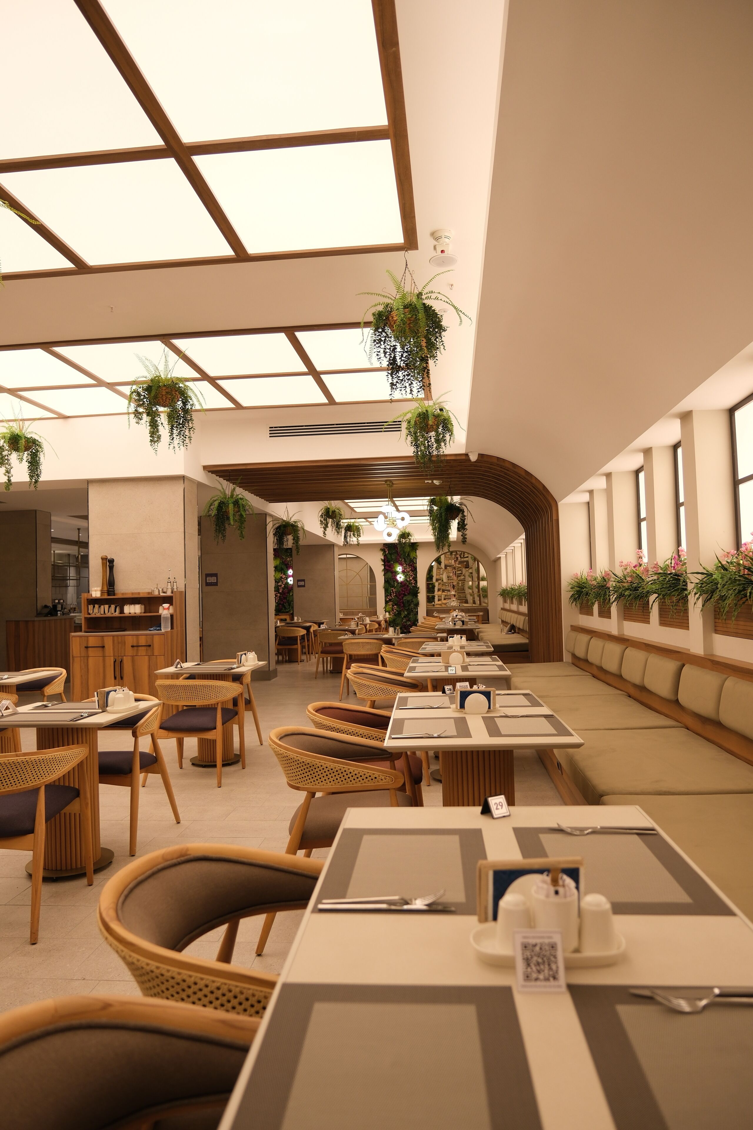 Een restaurant met houten tafels en stoelen, hangende planten en een hoge plafond met dakramen.