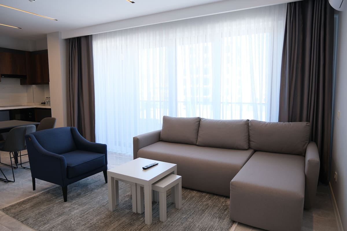 A modern living room with a sofa, armchair, and coffee table. There is a kitchen area in the background with a dining table and chairs.