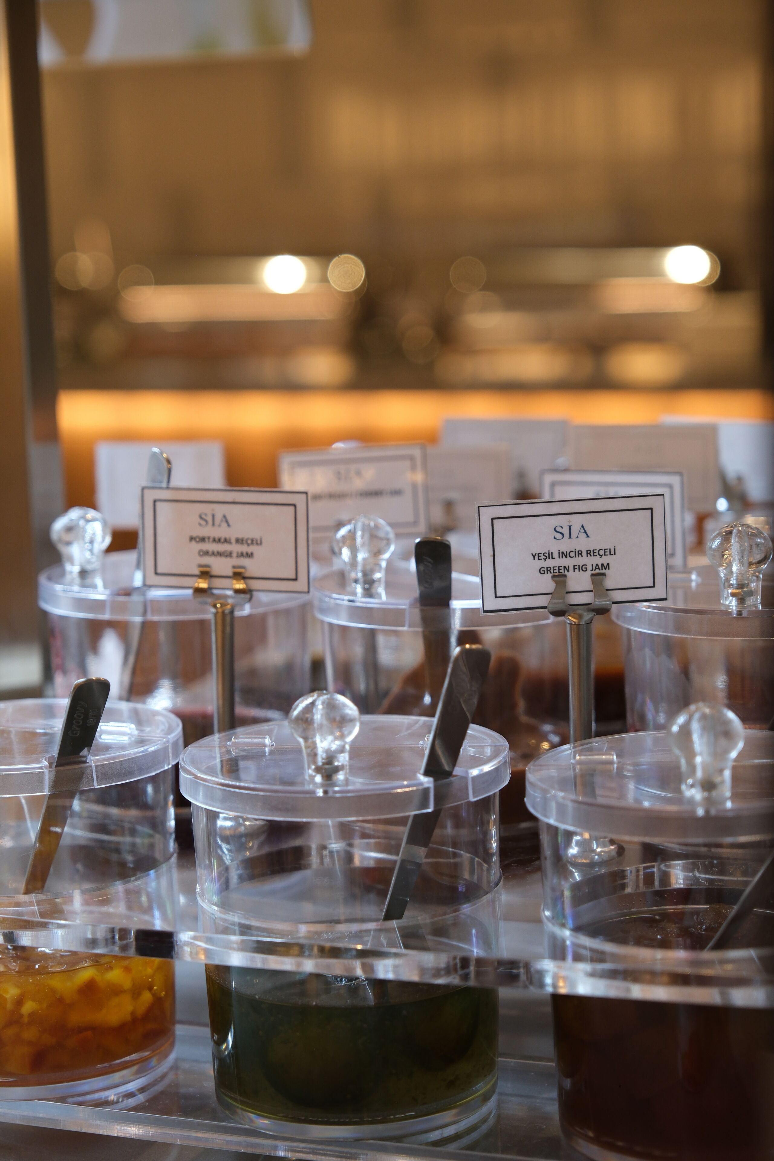 A selection of clear glass containers with metal spoons, each labeled with a name and type of jam.