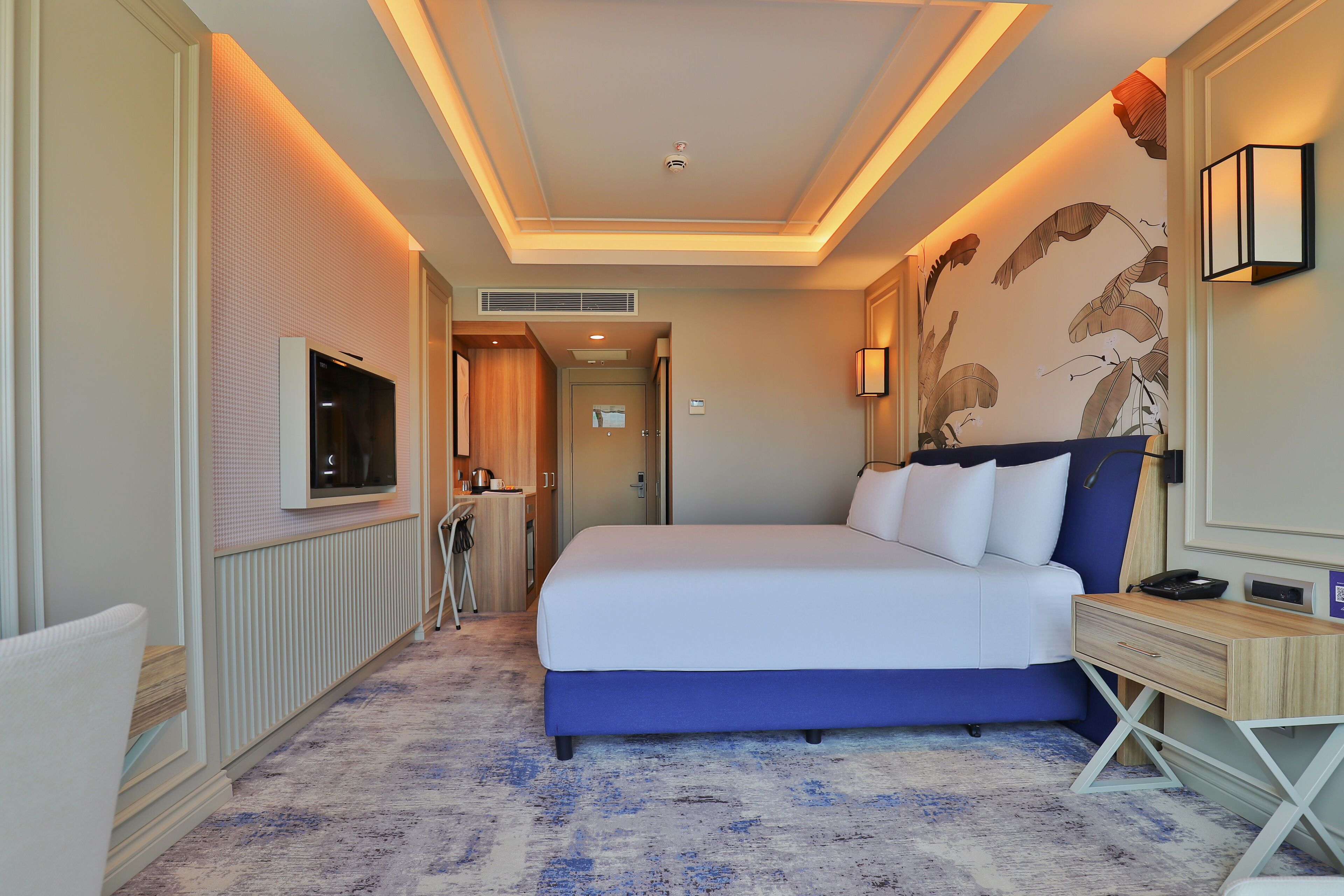 A hotel room with a large bed, a TV, a desk, and a wall with a floral mural.