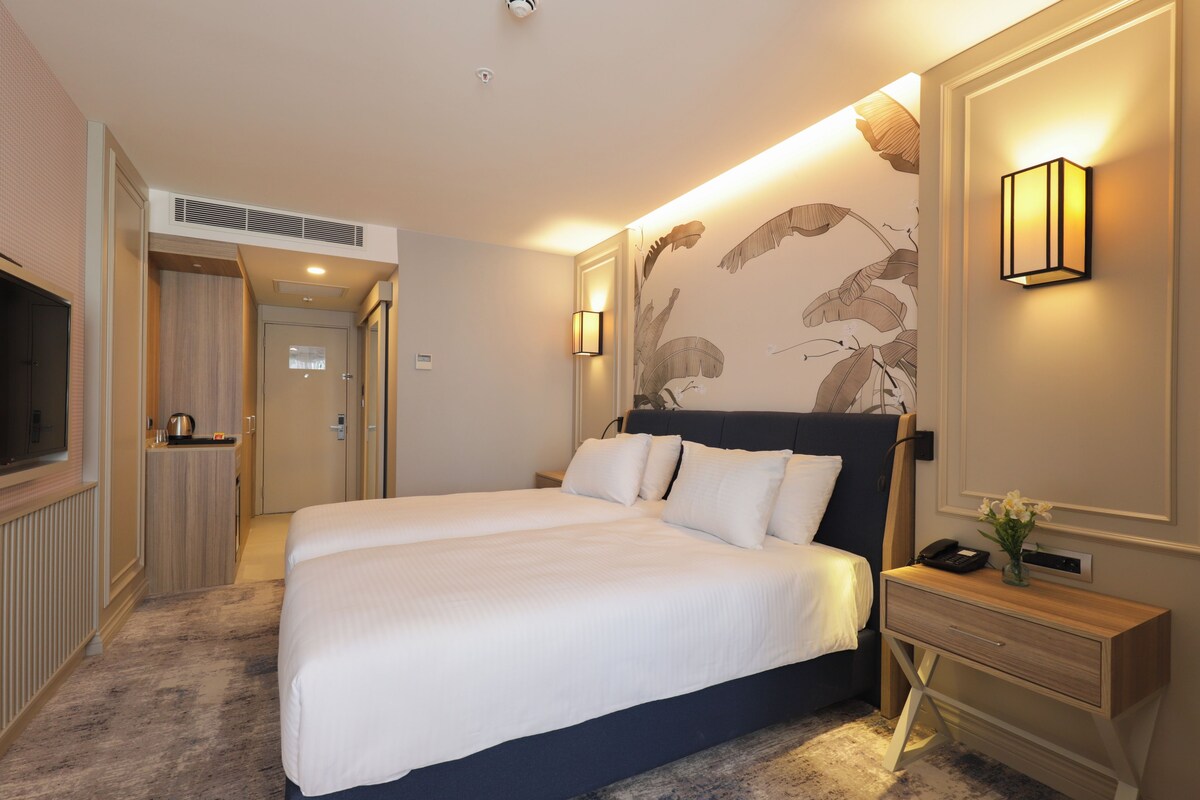 A hotel room with a large bed, a bedside table with a lamp, a TV, and a wall-mounted light fixture.