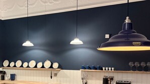 Una cucina con pareti blu scuro, pensili bianchi e un piano di lavoro in legno. Sono presenti tre lampade a sospensione, un lampadario grande e uno scaffale con piatti e tazze.