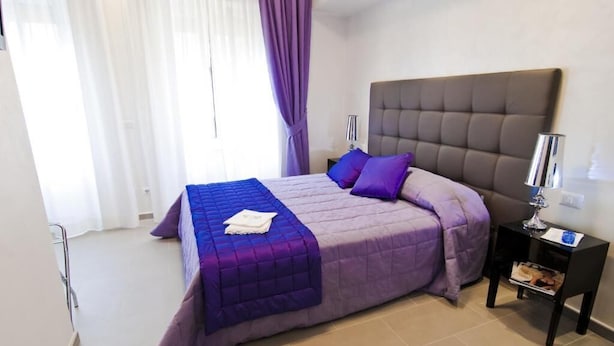 Une chambre d’hôtel moderne avec un grand lit, une literie violette, une tête de lit, une table de chevet avec une lampe et une fenêtre avec des rideaux.