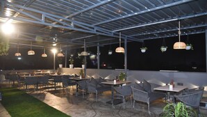 An outdoor patio area with tables and chairs, hanging plants, and a metal roof structure. - Aarchi International (Raipur)