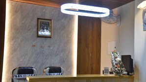 A reception area with a wooden reception desk, two chairs, a framed picture on the wall, and a ceiling light fixture. - Aarchi International (Raipur)