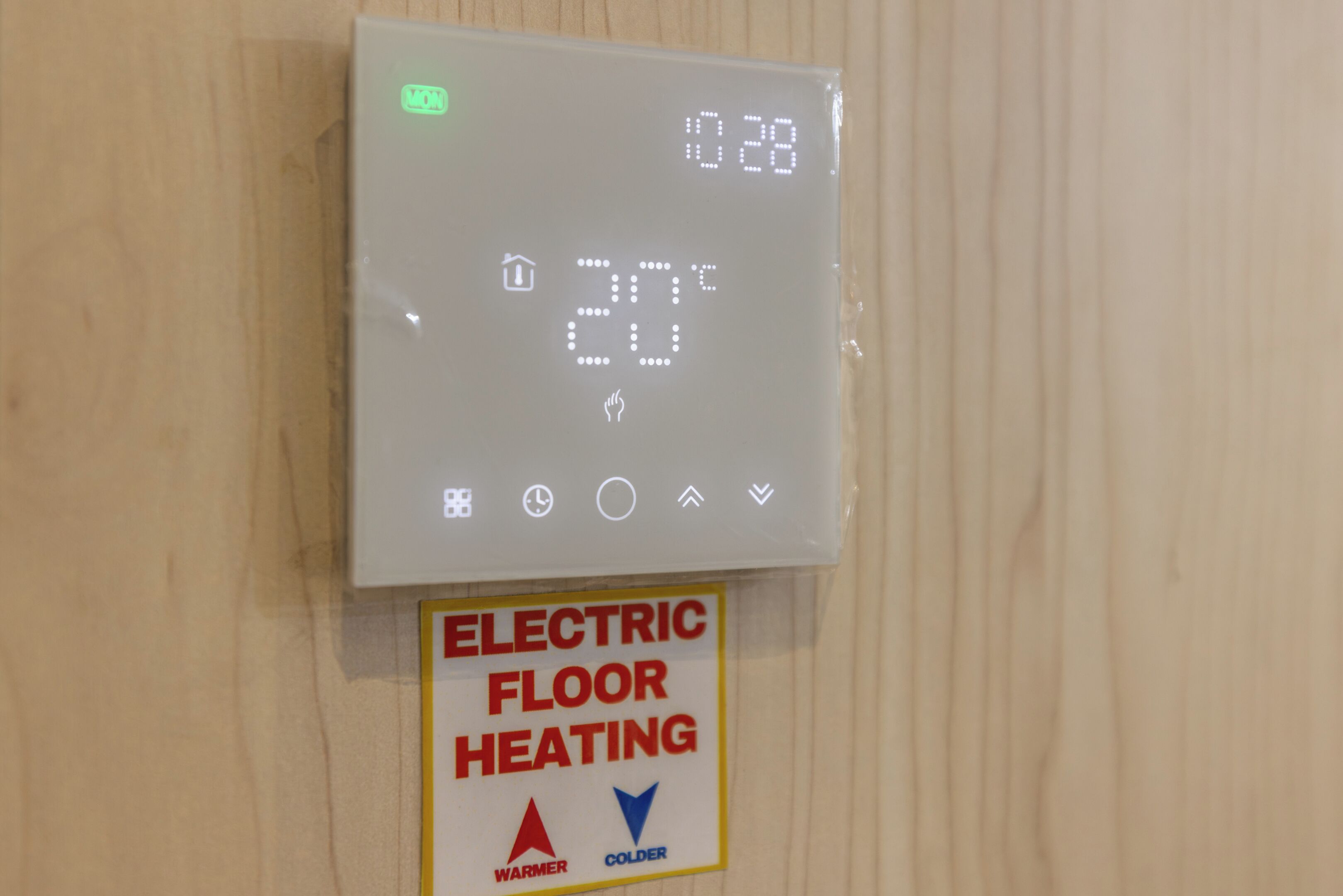 A digital thermostat with a display showing 20°C and a sign below stating "ELECTRIC FLOOR HEATING" with "WARMER" and "COLDER" buttons.