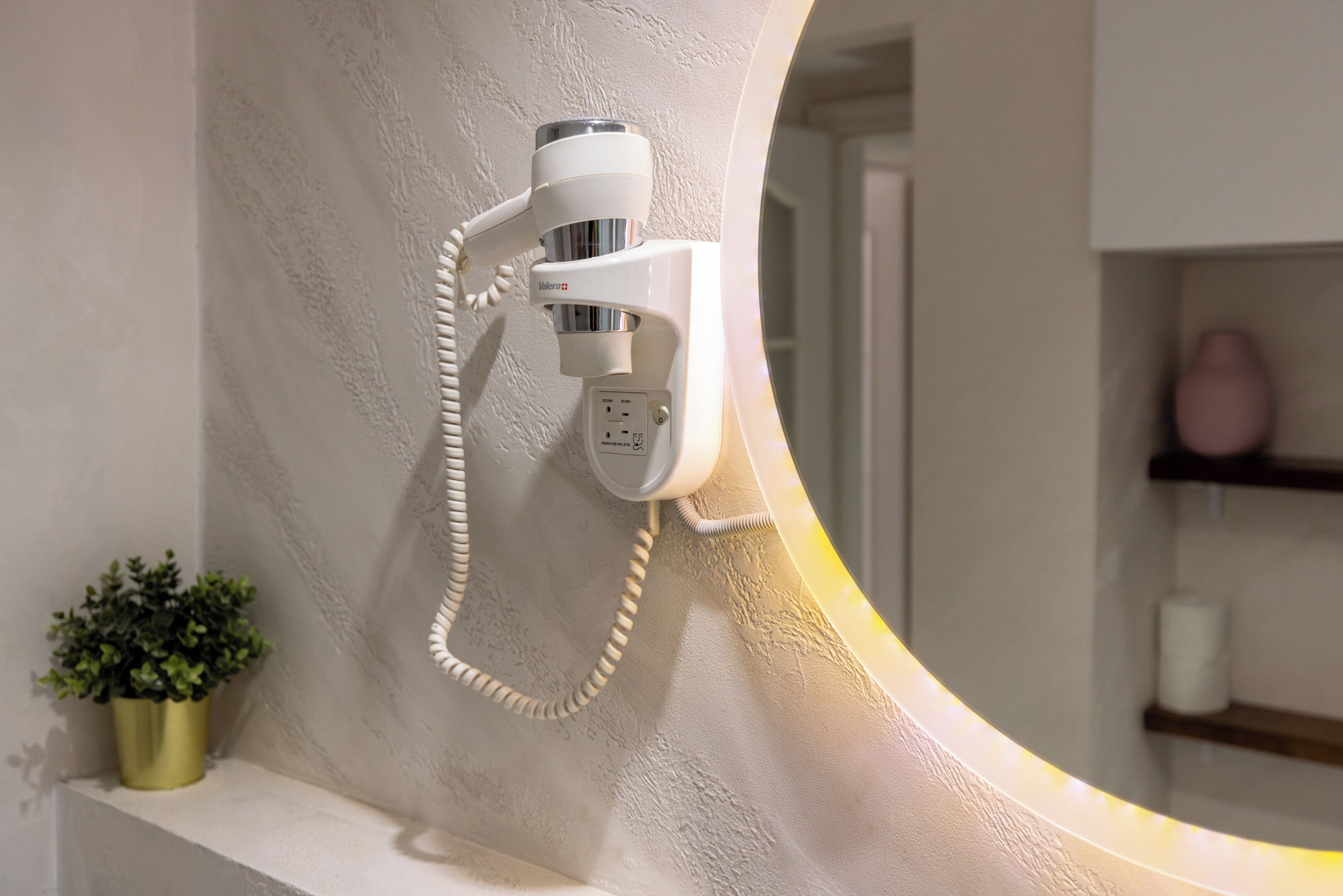 Un sèche-cheveux fixé au mur, avec un miroir qui reflète une étagère sur laquelle se trouvent un vase rose et un rouleau de papier toilette.