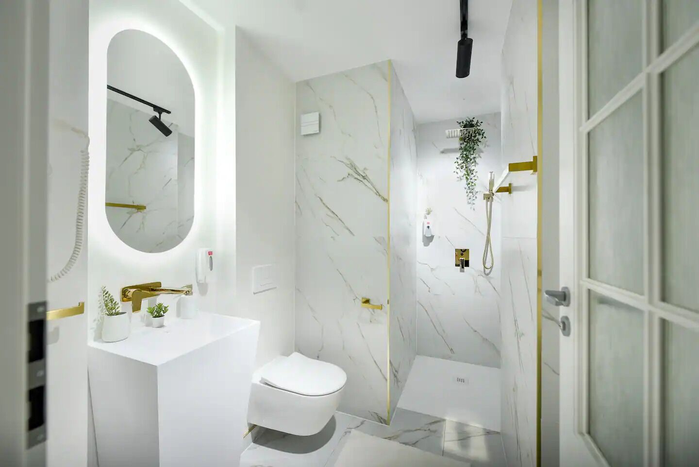 A modern bathroom with a white marble wall, a gold-framed sink, a toilet, and a shower with a gold faucet.