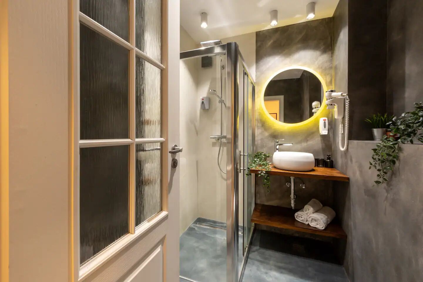 A modern bathroom with a glass shower enclosure, a round mirror with a yellow light, a white sink, and a wooden shelf with towels.