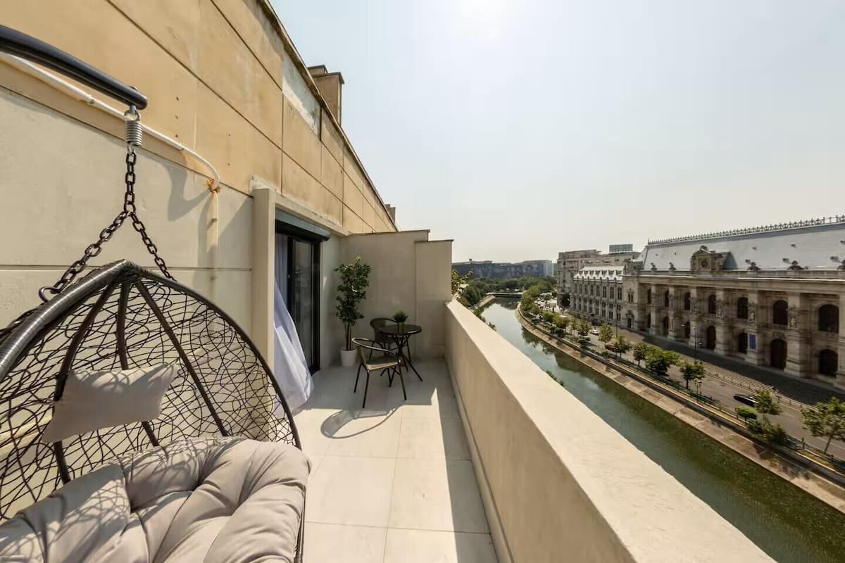 Un balcon agrémenté d’un fauteuil suspendu, d’une petite table avec des chaises, et offrant une vue sur une rivière et des bâtiments historiques.