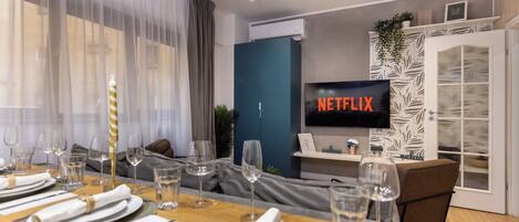 Ein modernes Wohnzimmer mit Essbereich, gedecktem Tisch und einer gemütlichen Sitzecke. Ein Fernseher zeigt 'NETFLIX'.