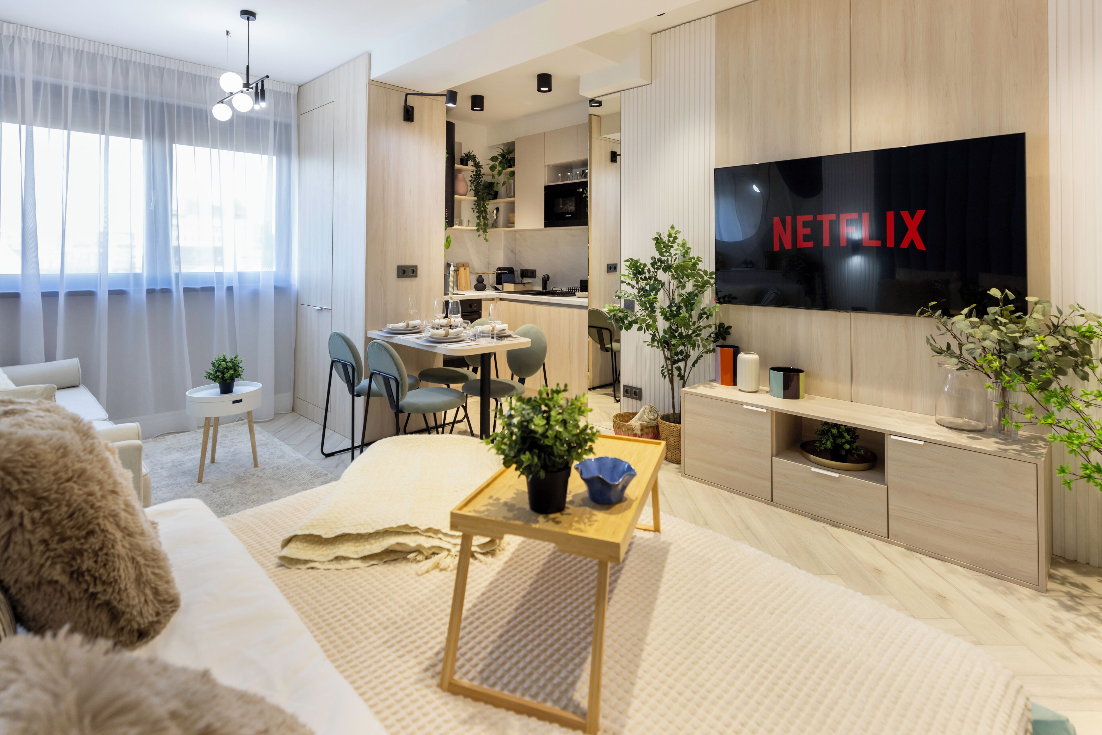 A modern living room with a large flat-screen TV displaying the Netflix logo, a dining area with chairs, and a kitchen visible through an open door.