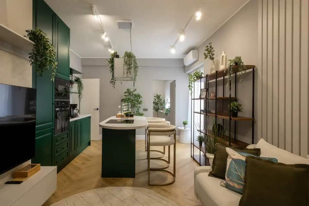 Une cuisine moderne avec des meubles de couleur verte, un îlot central et des plantes suspendues. Un canapé blanc avec des coussins et un téléviseur se trouvent à gauche.
