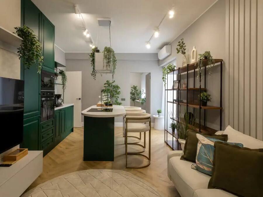 A modern kitchen with green cabinets, a central island, and hanging plants. There is a white sofa with cushions and a TV on the left.