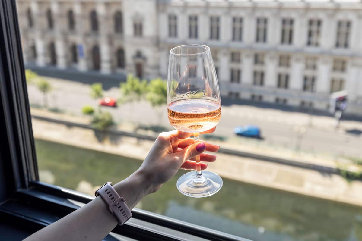 Une main tenant un verre à vin, avec en arrière-plan une rivière et des bâtiments.
