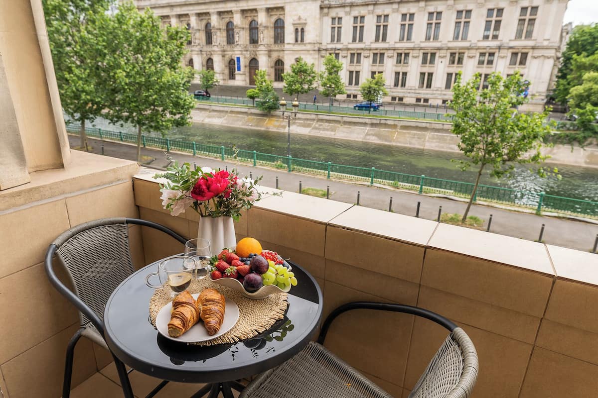 Un balcon avec une table préparée pour le petit-déjeuner, comprenant des croissants, des fruits et du café, donnant sur une rivière et des bâtiments historiques.