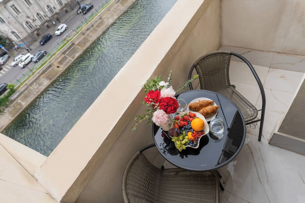 Un balcon avec une table dressée pour un repas, donnant sur un canal bordé d’immeubles et traversé par des voitures.