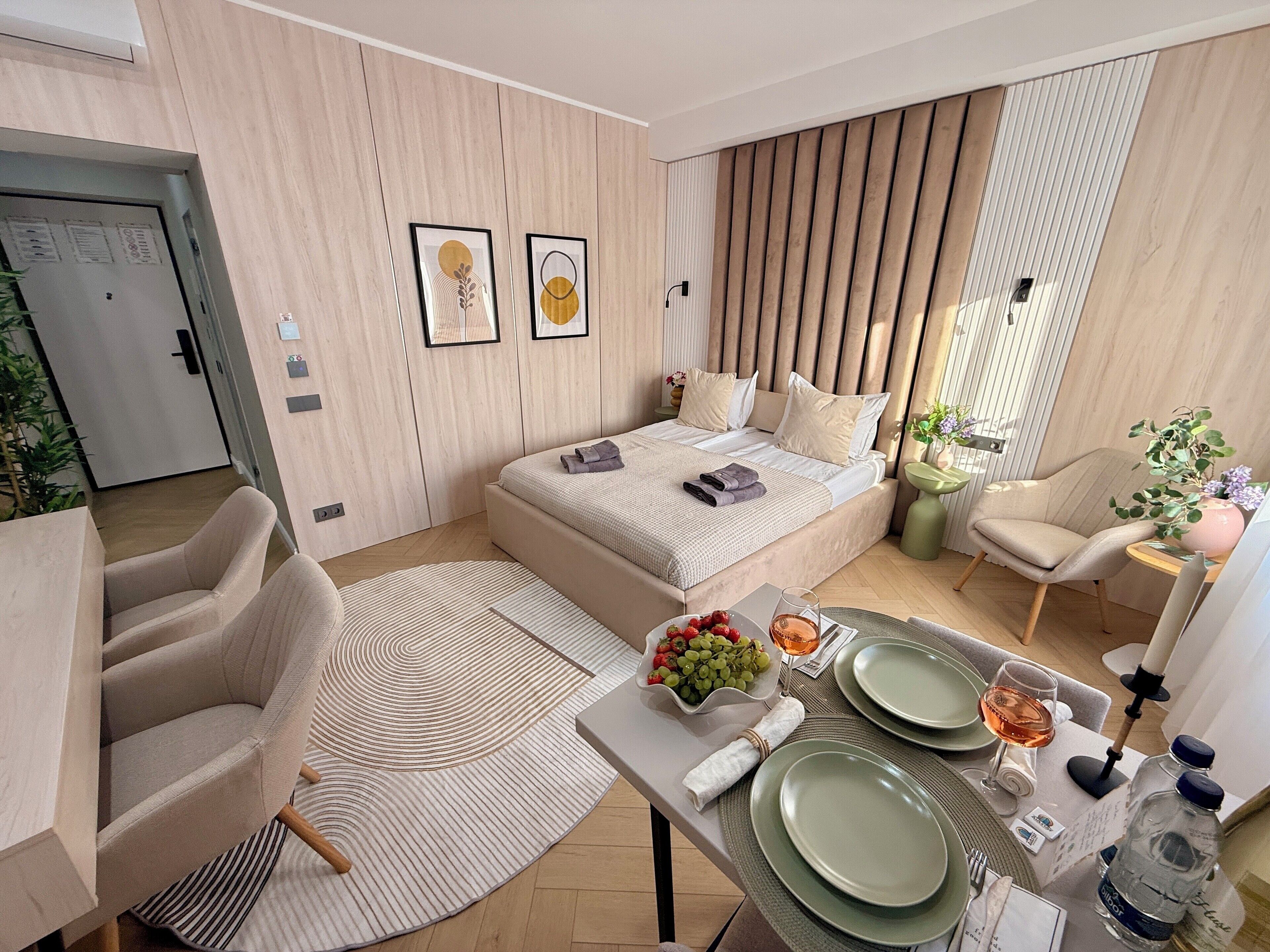 A modern bedroom with a large bed, a sitting area with a chair, a dining table set for two, and a view of the city through the window.