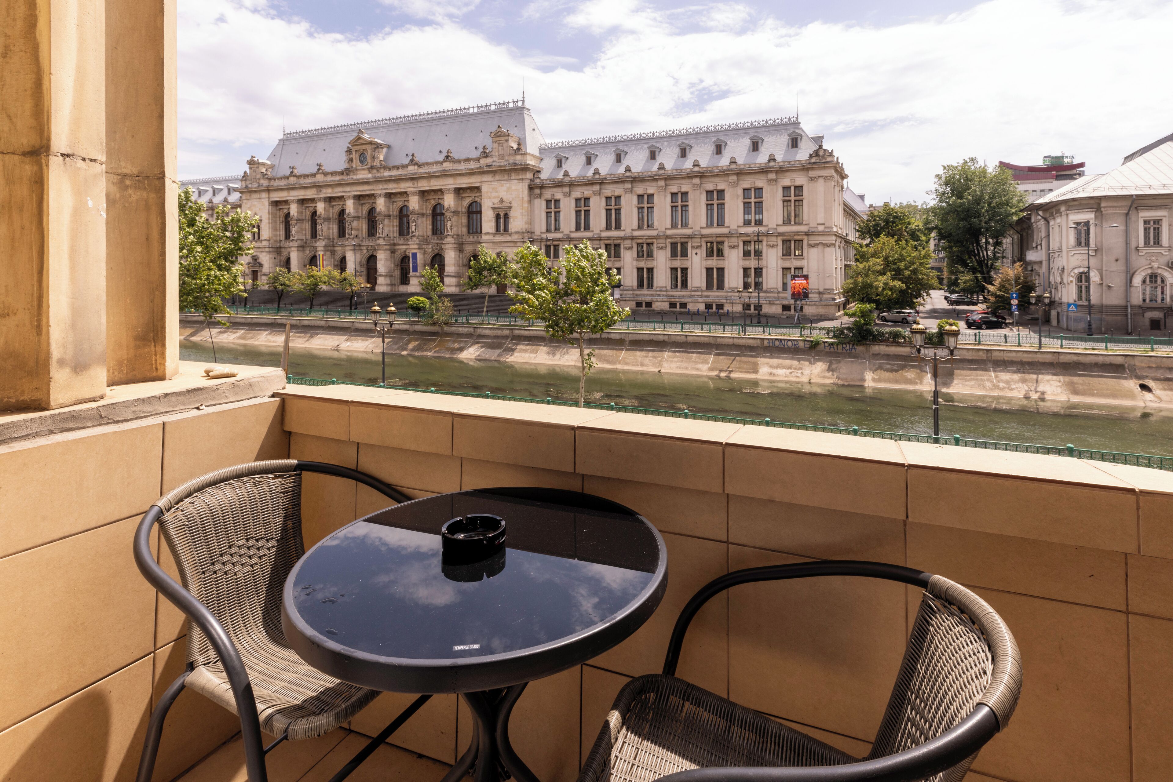 Een balkon met twee rieten stoelen en een kleine ronde tafel, met uitzicht op een rivier en historische gebouwen.