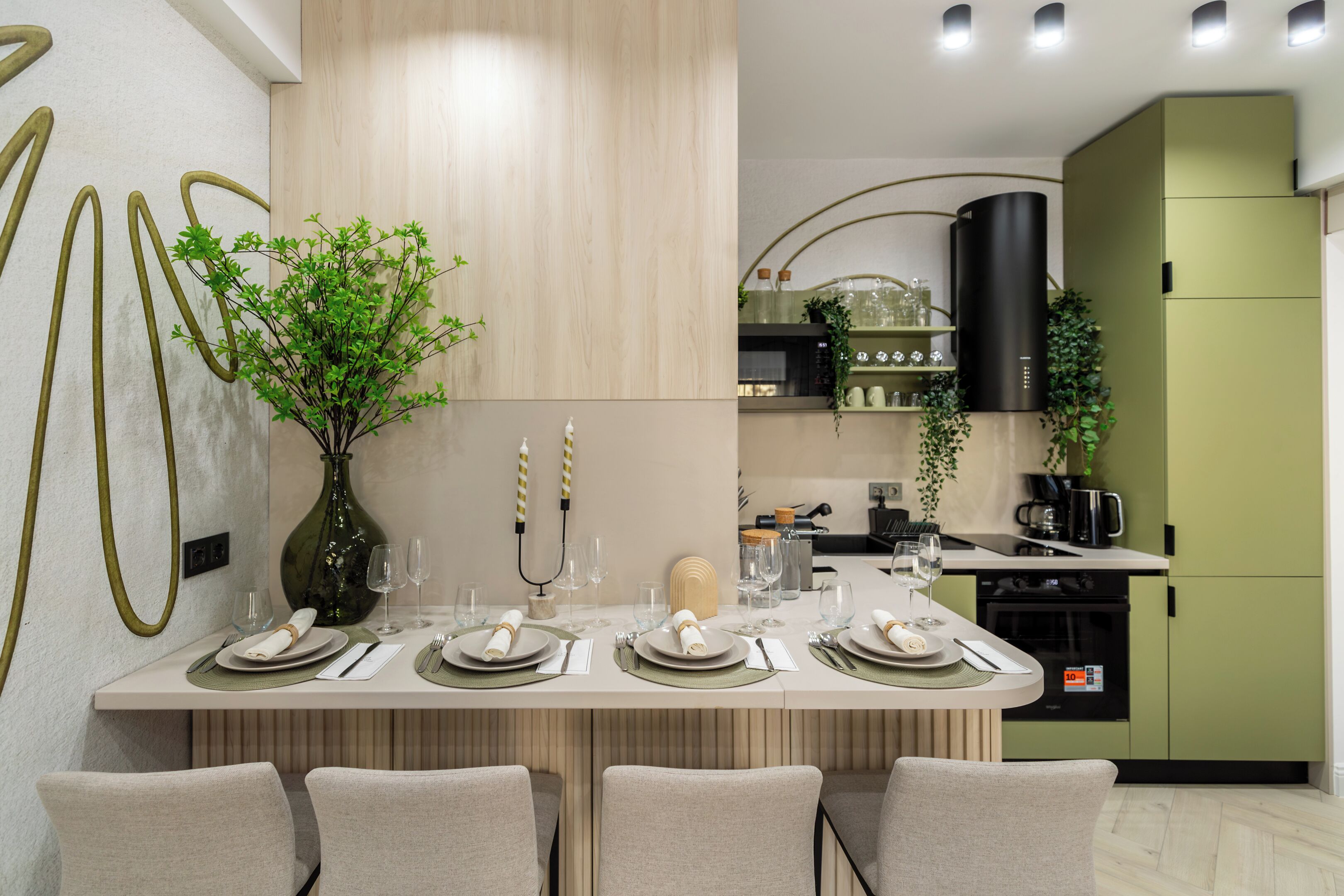 A modern dining area with a long table set for four, surrounded by chairs. A vase with green foliage is placed on the table, and there are various glassware and plates arranged neatly.