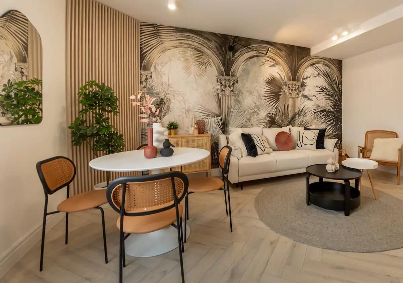 A modern living room with a round dining table, two chairs, a sofa with decorative pillows, a coffee table, and a large wall mural.