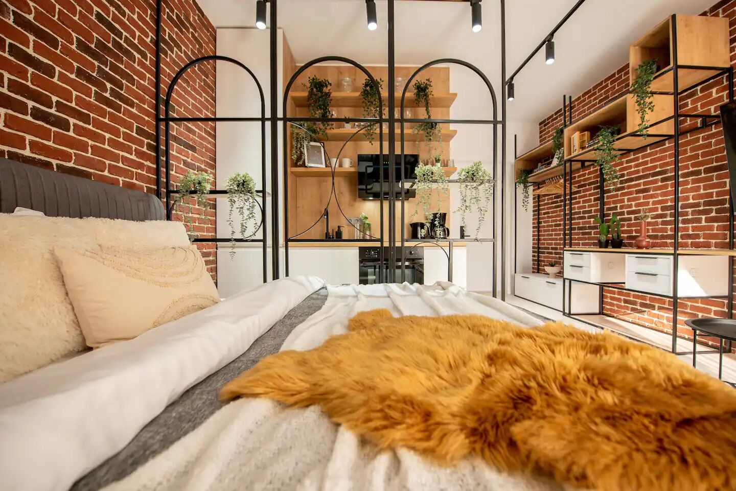 A modern bedroom with a brick wall, a bed with white bedding, a fur throw, and a kitchen area visible through a glass partition.