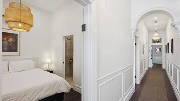 A hotel room with a bed, a nightstand, a lamp, a wicker chandelier, a door leading to a bathroom, and a hallway with a carpet and framed pictures.