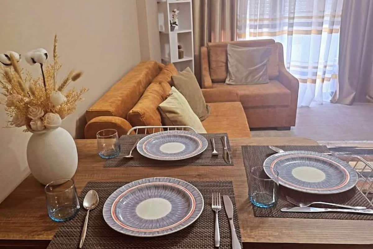 A dining area with a table set for four, a sofa, a bookshelf, and a painting on the wall.