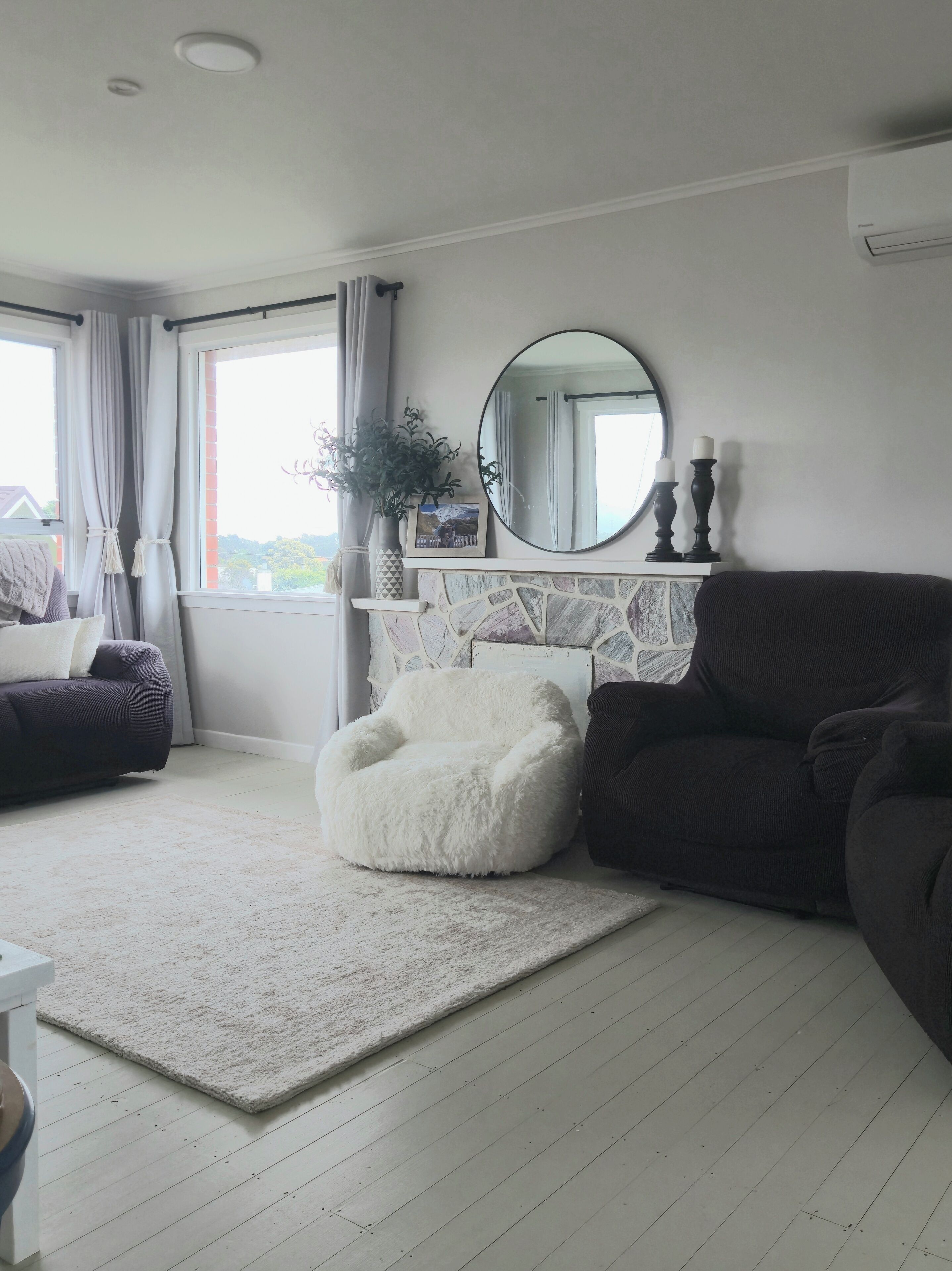A living room with a white fluffy chair, dark sofas, a white rug, a fireplace with a stone mantle, a round mirror, and a window with curtains.