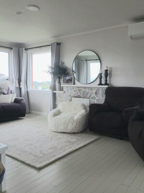 A living room with a white fluffy chair, dark sofas, a white rug, a fireplace with a stone mantle, a round mirror, and a window with curtains.