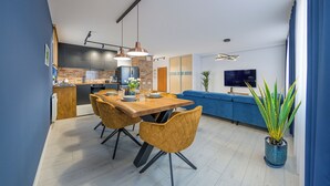 Una cocina moderna con un salpicadero de ladrillo, una zona de comedor con una mesa y sillas de madera, y un salón con un sofá azul y una televisión.