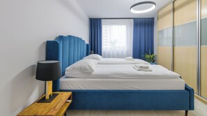 Una habitación de hotel moderna con una cama grande, una mesita de noche con lámpara, un armario y una ventana con cortinas azules.