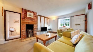 A cozy living room with a brick fireplace, a sofa, a coffee table, and a television. - Countryside Cottage in Bulmer- Sleeps 6/7 (Sudbury)