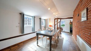 A dining area with a table, chairs, and a patterned rug. There are windows with curtains, a brick wall, and a ceiling with exposed beams. - Countryside Cottage in Bulmer- Sleeps 6/7 (Sudbury)