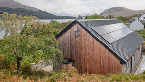 A wooden cabin with a sloped roof, a small window, and a door, surrounded by natural vegetation and a lake view.