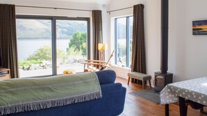 A room with a large window overlooking a lake, a sofa, a table with a polka dot tablecloth, and a wood-burning stove.