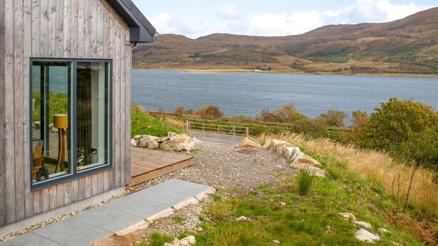 A modern wooden building with large windows overlooking a lake and hills.
