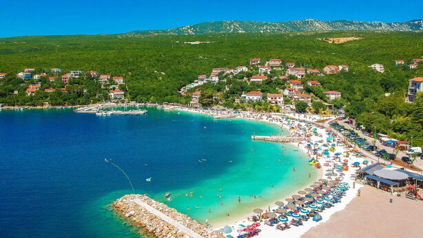 A coastal town with a beach, clear blue waters, and houses on the hillside.