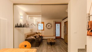 Ein modernes Wohnzimmer mit einem Holzesstisch, einer Couch und einem Couchtisch. An der Wand hängt eine Uhr, und eine Tür führt in einen weiteren Raum.