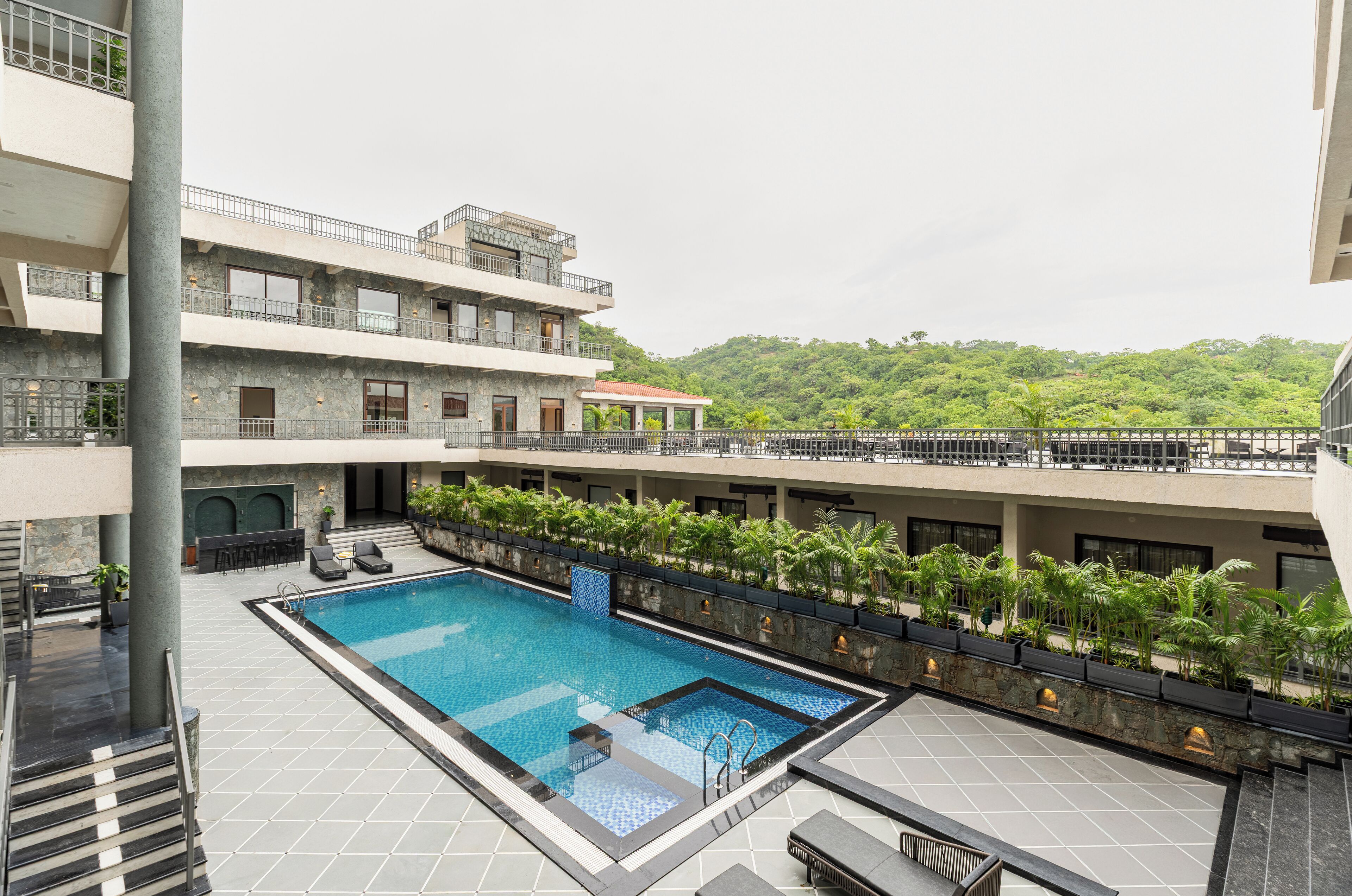 A rooftop swimming pool with multiple levels of balconies, surrounded by lush greenery and a mountainous backdrop.